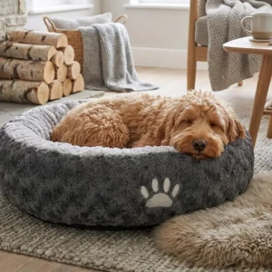 Ein flauschiger, hellbrauner Hund schläft in einem Nobby Donut SEOLI - Hundebett mit Pfotenabdruck, umgeben von kuscheligen Decken, Kaminholz und sanftem Tageslicht in einem stilvollen Wohnzimmer.